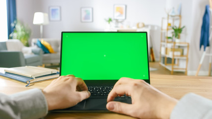 First Person Close-up Shot Man Uses Laptop with Green Mock-up Screen While Sitting at the Desk in...