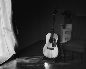 Fototapeta na wymiar Black & white photo of acoustic Spanish guitar on a stand in the moody shadows of a dark room with bright light coming in from behind a curtain