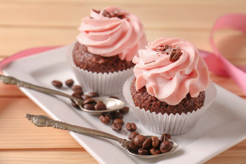Plate with sweet cupcakes, coffee beans and spoons on wooden table, closeup