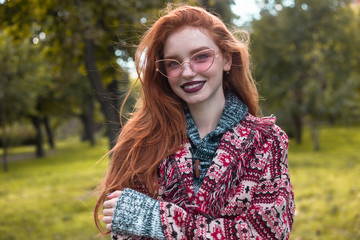 Naklejka premium Redhead young happy woman with freckles and dark lipstick wearing sunglasses posing outdoors in park. Looking camera.