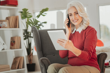 Pleasant conversation. Smiling senior lady in spacious living room holding tablet while sitting in chair and talking on the phone