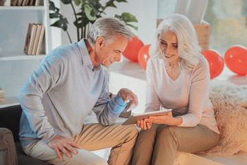 Obraz premium Valuable memories. Charming elderly woman siting in living room with a tablet and showing pictures to her pleased positive husband sitting next to her