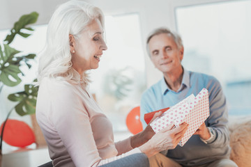 Many thanks. Smiling elderly lady enjoying her present and being pleased with long-awaited surprise inside the present while happy attentive husband looking at her