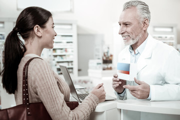 Obraz na płótnie Canvas Recommending pills. Pleasant smiling bearded pharmacist wearing white coat recommending good pills his visitor