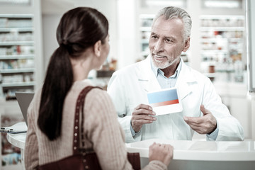 Fototapeta premium Pill box. Bearded mature pharmacist wearing white coat holding pill box while providing service his client