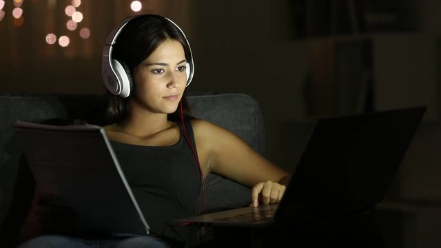 Serious teenage student comparing notes with a laptop sitting on a couch in the night at home