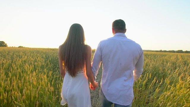 Happy young couple holding hands and walking through wheat field at summer sunset, having fun outdoors. Love, summer time. Man and woman, happy family walking on meadow. Nature, Sun flare Slow motion