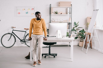 Fototapeta na wymiar handsome african american designer in orange sweater leaning on table and looking away in office