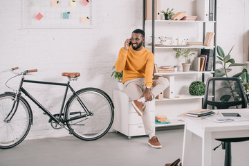 smiling handsome african american designer in orange sweater talking by smartphone in office