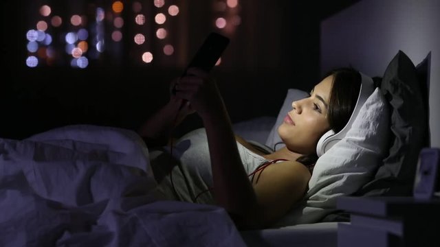 Serious girl with headphones browsing media in a phone lying on a bed in the night at home