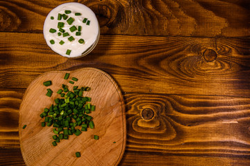 Glass bowl with sour cream and cutting board with chopped green onion on wooden table. Top view