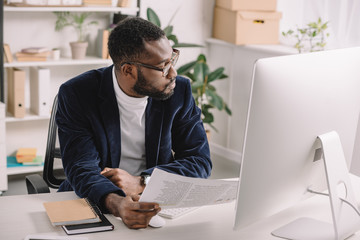 Naklejka premium bearded african american businessman doing paperwork in office with computer