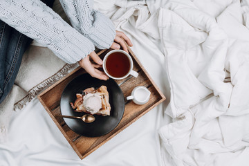Cozy flatlay of bed with wooden tray with pie, ice cream and black tea and woman's hands in grey sweater holding cup