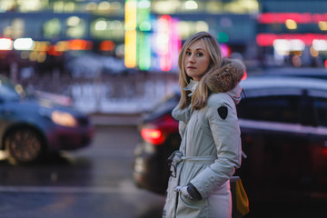 Beautiful young woman with big eyes and long blond hair in jacket with hood stands on evening city street.