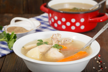 Hot homemade chicken soup in white bowl - alternative remedy for cold and flu. Red pot with soup in background on wooden table.