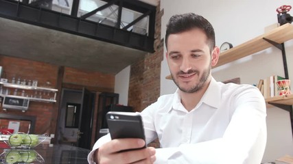 Joyful good-looking businessman in white shirt using smartphone, smiling. Fabulous young man using device. Indoors. Apartment. Daytime.
