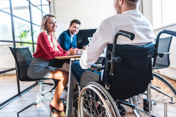partial view of smiling businesspeople looking at colleague in wheelchair in office