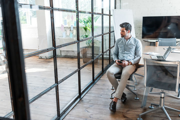 Fototapeta premium high angle view of young businessman using smartphone and looking away while sitting in office
