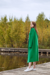 Girl in a green coat walks along the lake embankment on a cloudy autumn day. Autumn fashion and clothing, yellow fallen leaves floating in the water. Romantic mood