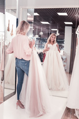 Fototapeta premium The most beautiful bride. Joyful delighted woman looking at the wedding dress while standing in front of the mirror