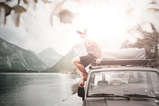 mann mit hut und tank-top sitzt auf dem dachtr&auml;ger eines gel&auml;ndewagens an einem see mit hellen sonnenstahlen und bilckt auf die Tiroler Alpen
