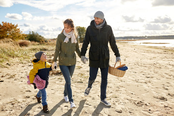 family, leisure and people concept - happy mother, father and little son walking along autumn beach with picnic basket and blanket