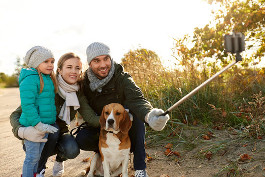 family, pets and people concept - happy mother, father and little daughter with beagle dog taking picture by smartphone on selfie stick outdoors in autumn