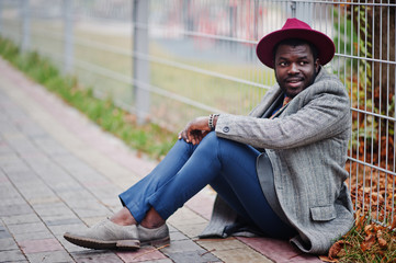 Stylish African American man model in gray coat, jacket tie and red hat sitting.
