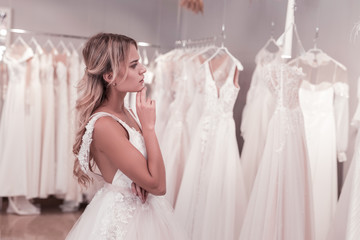 Which one is best. Pleasant thoughtful bride looking at the dresses while thinking which one to...