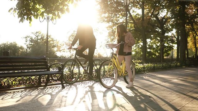 Beautiful young couple riding their bikes in the empty city park or boulevard in summertime. Stop and sit on the bench and embracing. Loving couple. Sun shines on the background