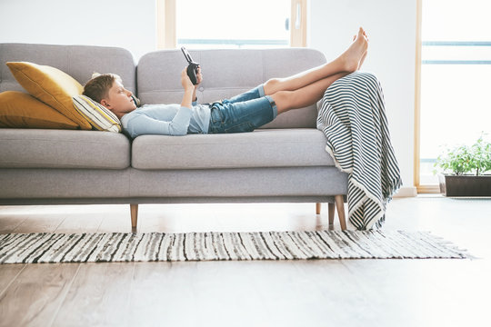 Boy plays with electronic devices - gamepad connected with smartphone lying on the cozy sofa in the home living room.