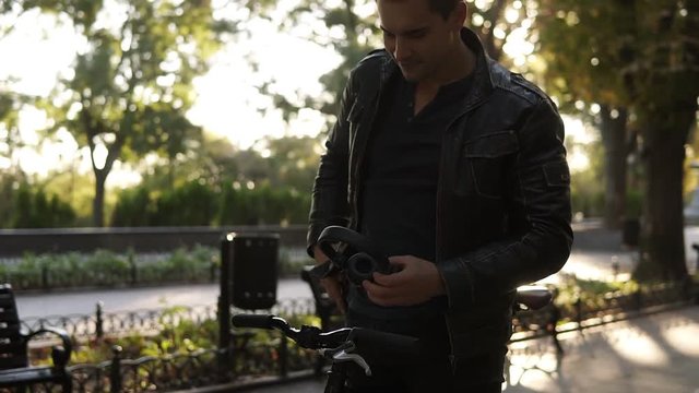 Lifestyle, transport, technology and people concept - young man with headphones starts riding bicycle on city street, choosing the favourite mucis on his smartphone. Autumn in the city park