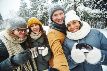 Positive excited young multi-ethnic friends in warm clothing holding thermos mugs and taking selfie...