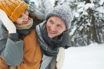 Smiling handsome young man in knitted hat and scarf giving girlfriend piggyback ride in winter forest, she adjusting hat