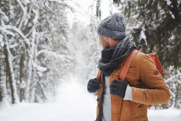 Fototapeta na wymiar Pensive young male backpacker in knitted hat and scarf holding handles of backpack and looking back in forest while contemplating winter nature