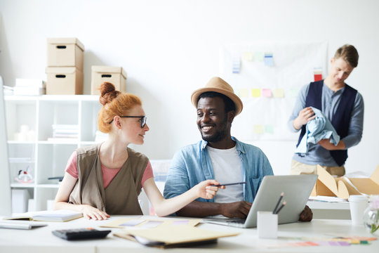 Young woman discussing with her African colleague presentation on laptop and male designer packing clothes in the background