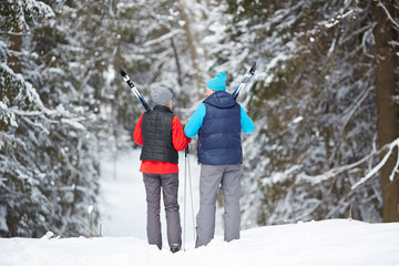 Fototapeta na wymiar Rear view of sporty aged couple with skiing equipment walking down forest path on winter day