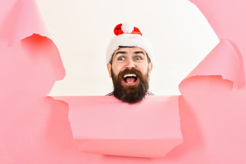 Bearded man in santa hat looking through hole in paper. Christmas, New Year, holidays, winter concept. Stylish guy in santa hat. Smiling guy in santa hat breaks through paper wall. Christmas sales.
