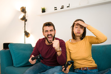 Beautiful couple playing video games at home, sitting on a cozy couch, the guy is laughing and squeezing fist because he is the winner and girl is making angry faces.