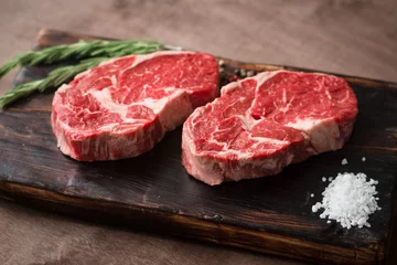  Twee verse rauwe rib-eye steak op een houten bord op een houten ondergrond met zout, peper en rozemarijn in een rustieke stijl © Andrey