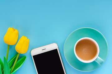 Smartphone with cup of coffee and artificial yellow tulips on blue background for media technology and modern lifestyles concept