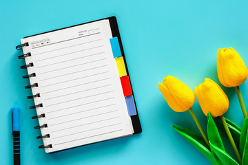 Blank notebook and a pen with artificial yellow tulips on blue background for workspace, nature decoration and springtime concept