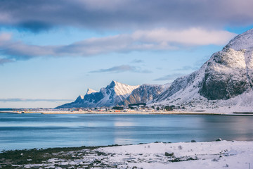 Lofoten Islands in winter. Beautiful daytime landscape with snowy rocky mountains, blue cloudy sky and small village on the sea coast, Northern Norway