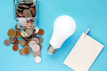 Coins in glass jar and outside, Thai currency money with glowing LED light bulb and blank white paper card on blue background for energy savings and planning concept