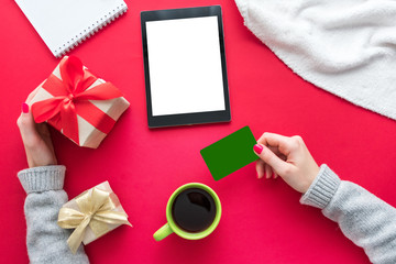 Obraz premium Female hands, woman holding a Business card and gift. Cup of coffee or tea, tablet pc, red table, gifts boxes for the holidays, background with copy space for advertisement, top view