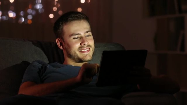 Man watching media with a tablet in the night sitting on a couch in the living room at home