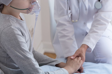 Closeup of sick kid with oxygen mask and doctor holding her hand in hospital