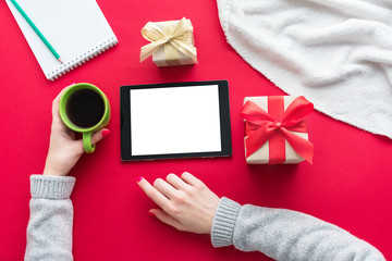 Female hands, woman holding a Cup of coffee or tea, girl uses the tablet pc. Red table, gifts boxes for the holidays, background with copy space for advertisement, top view