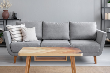 Wooden table and big grey couch with pillows in living room of trendy apartment, real photo with...