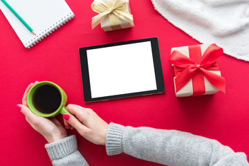 Obraz premium Female hands, woman holding a Cup of coffee or tea. Tablet pc, red table, gifts boxes for the holidays, background with copy space for advertisement, top view